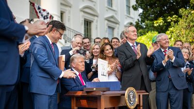 A press photo of President Donald Trump sitting at a desk holding a signed document, surrounded by applauding people in formal attire outside the White House.